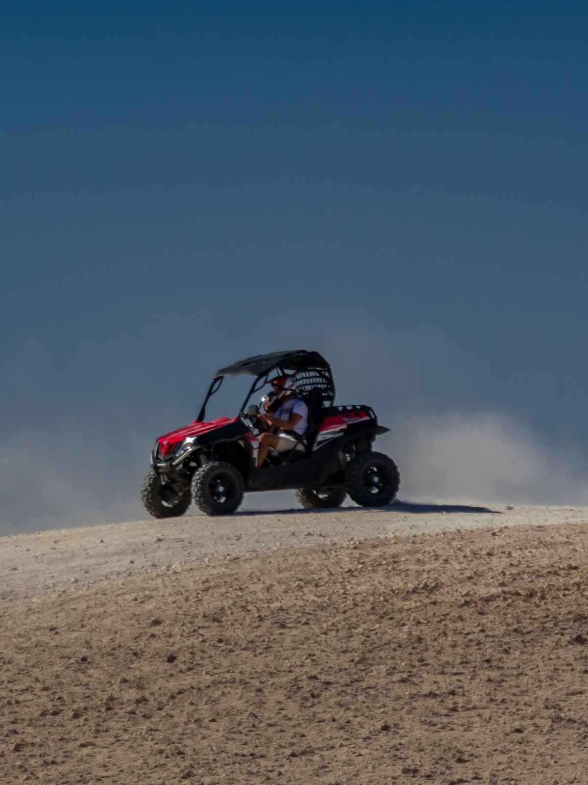 Red and black buggy cresting a ridge, helmeted driver, a cloud of dust behind