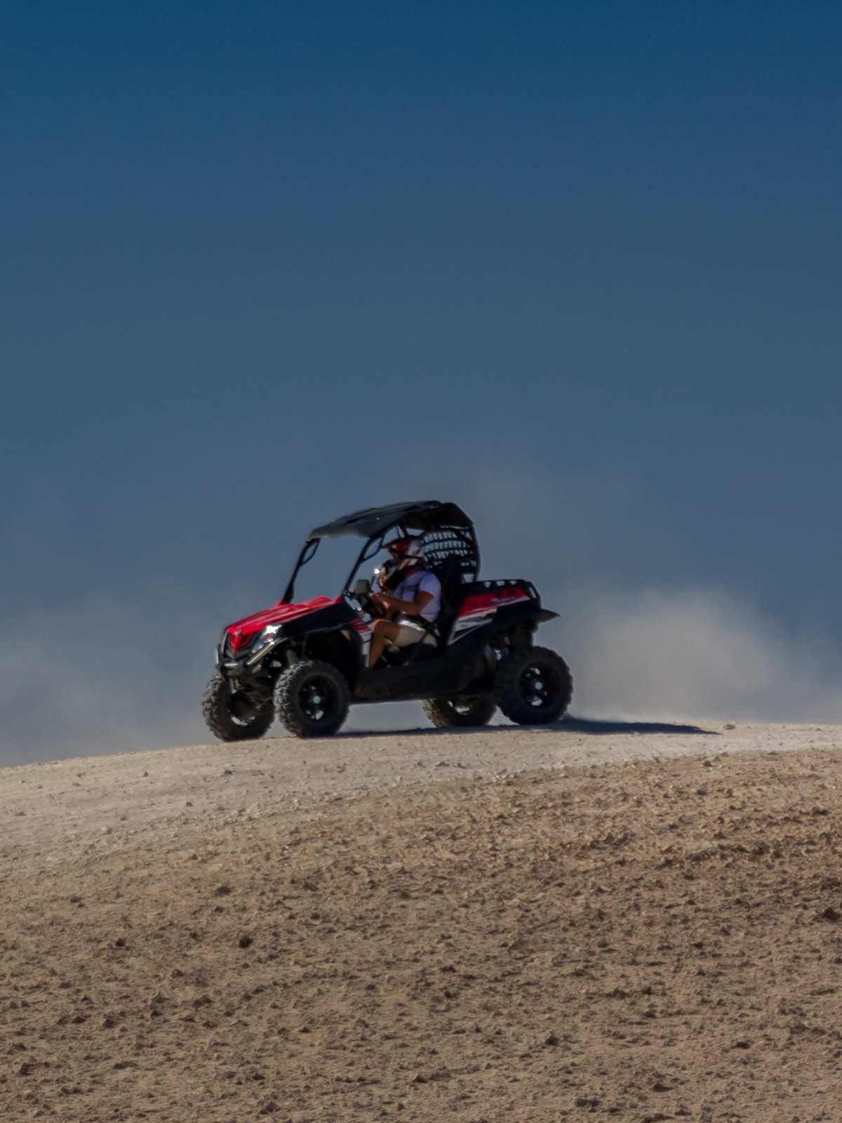 Rot-schwarzer Buggy auf einer Hügelkuppe, Fahrer mit Helm, dahinter eine Staubwolke