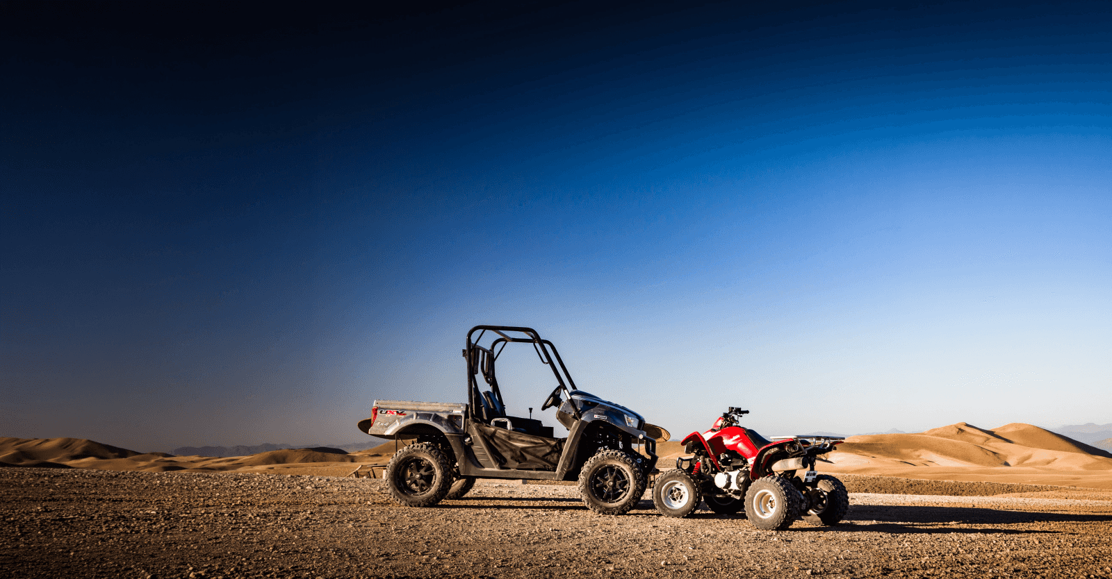 Ein Buggy und ein rotes Quad nebeneinander auf einer steinigen Ebene, Sandhügel am Horizont unter tiefblauem Himmel