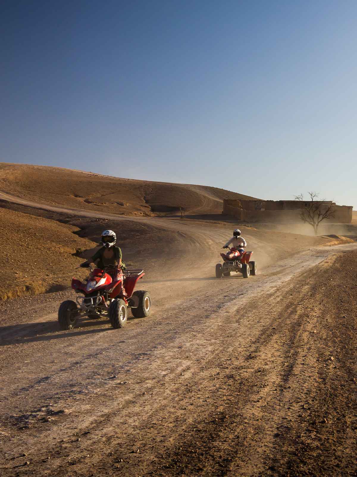 Two riders on red quad bikes on a dusty track, hills and a mud-brick building behind