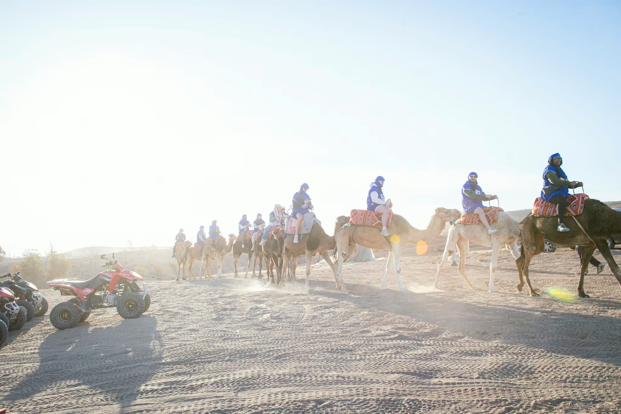 Kamelkarawane im Gegenlicht und Staub, seitlich geparkte Quads