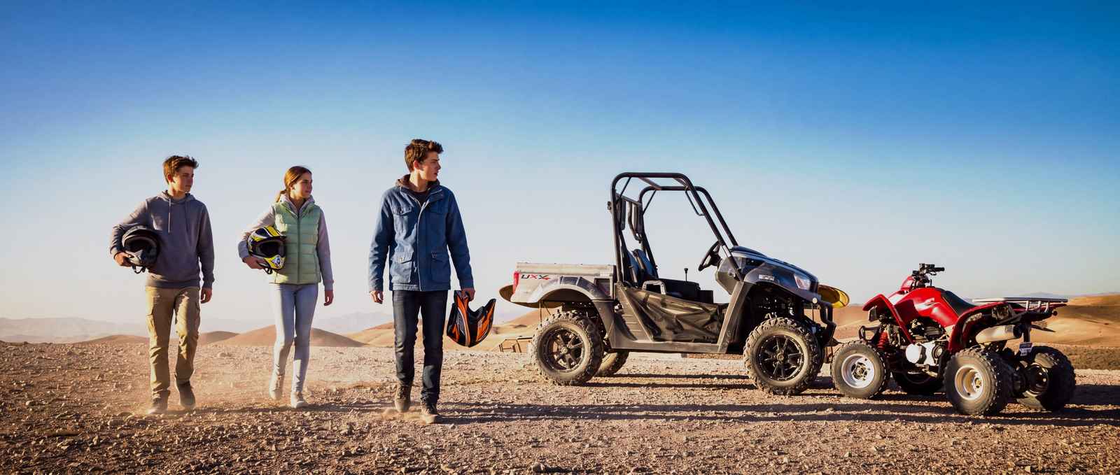 Three young riders, helmets in hand, walking on a stony track beside a buggy and a red quad, hills in the background