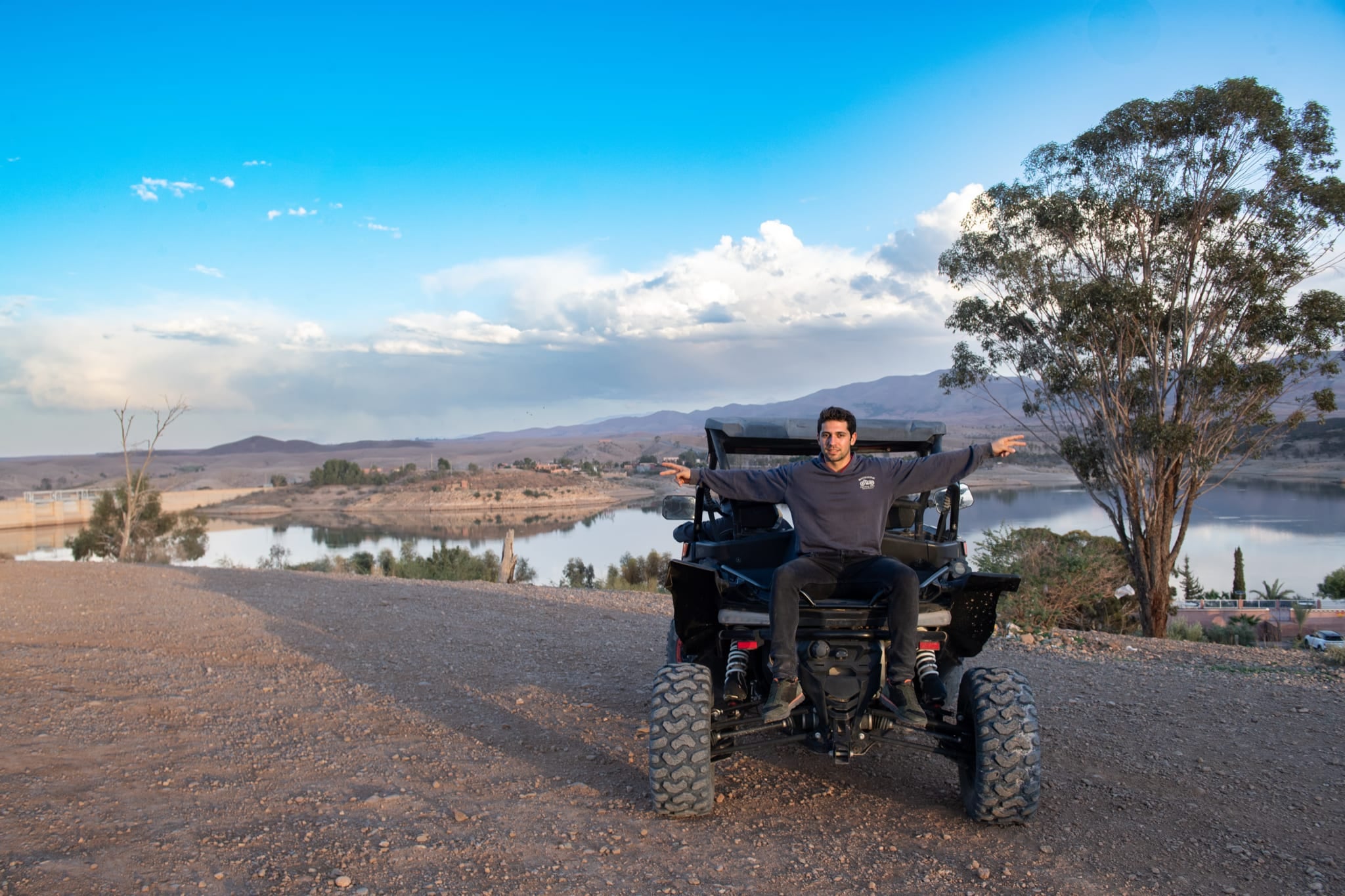 Mann sitzt mit ausgebreiteten Armen hinten auf einem Buggy, über dem Stausee Lalla Takerkoust