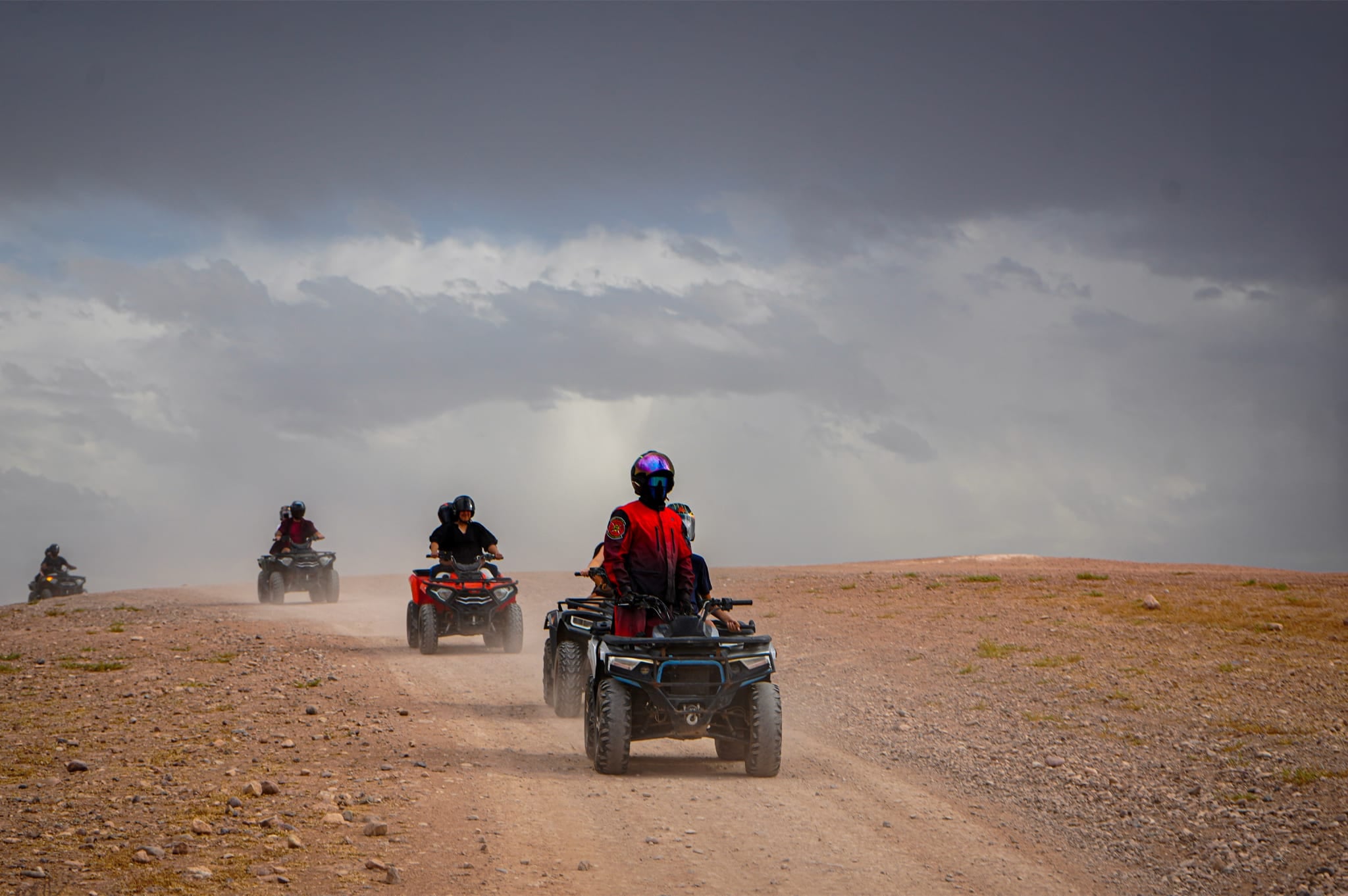 Eine Reihe Quads auf einer staubigen Piste unter Gewitterhimmel, vorne ein Fahrer in roter Jacke