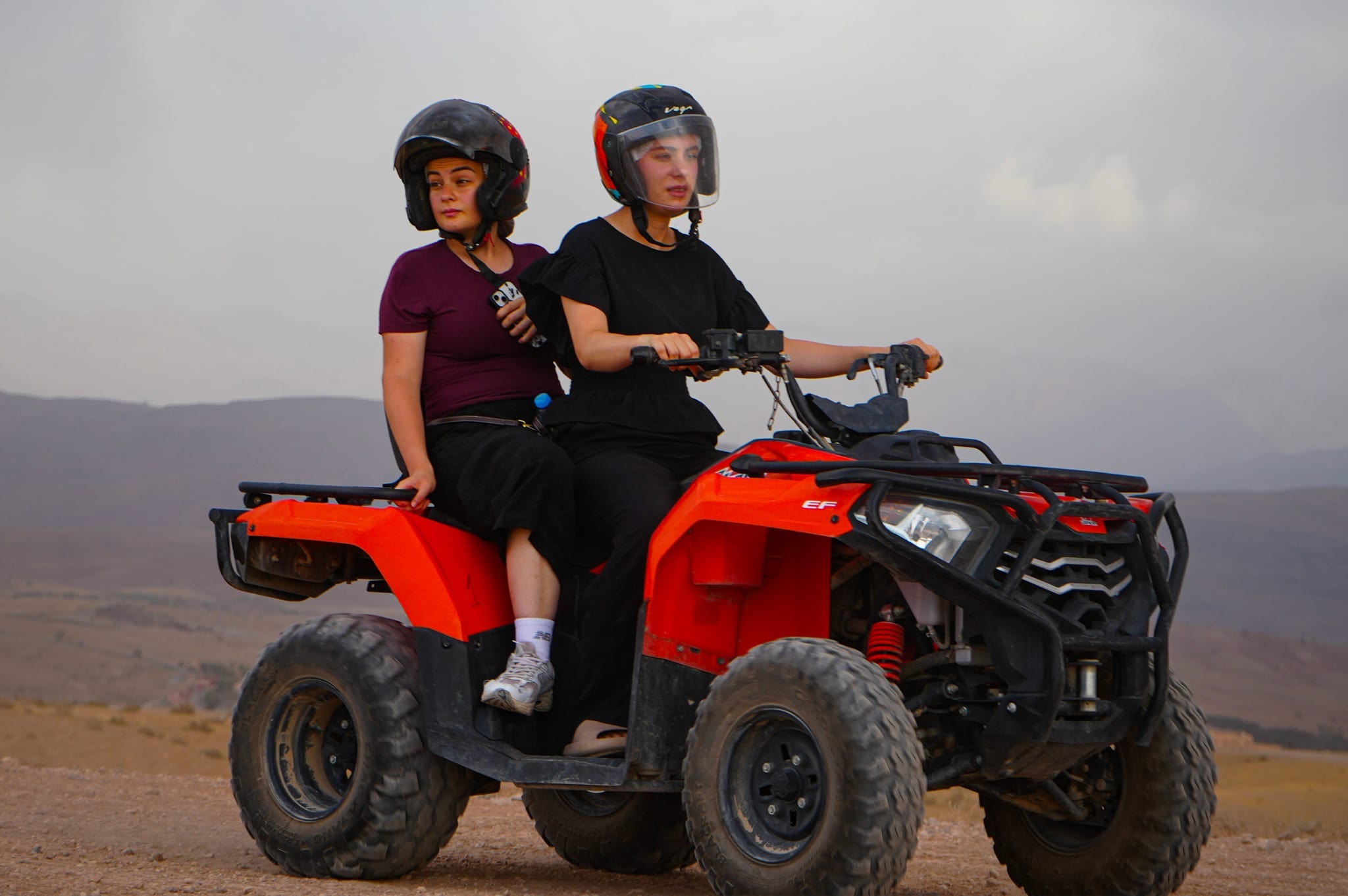 Zwei junge Frauen mit Helm auf einem roten Quad, eine am Lenker, dahinter Hügel im Dunst