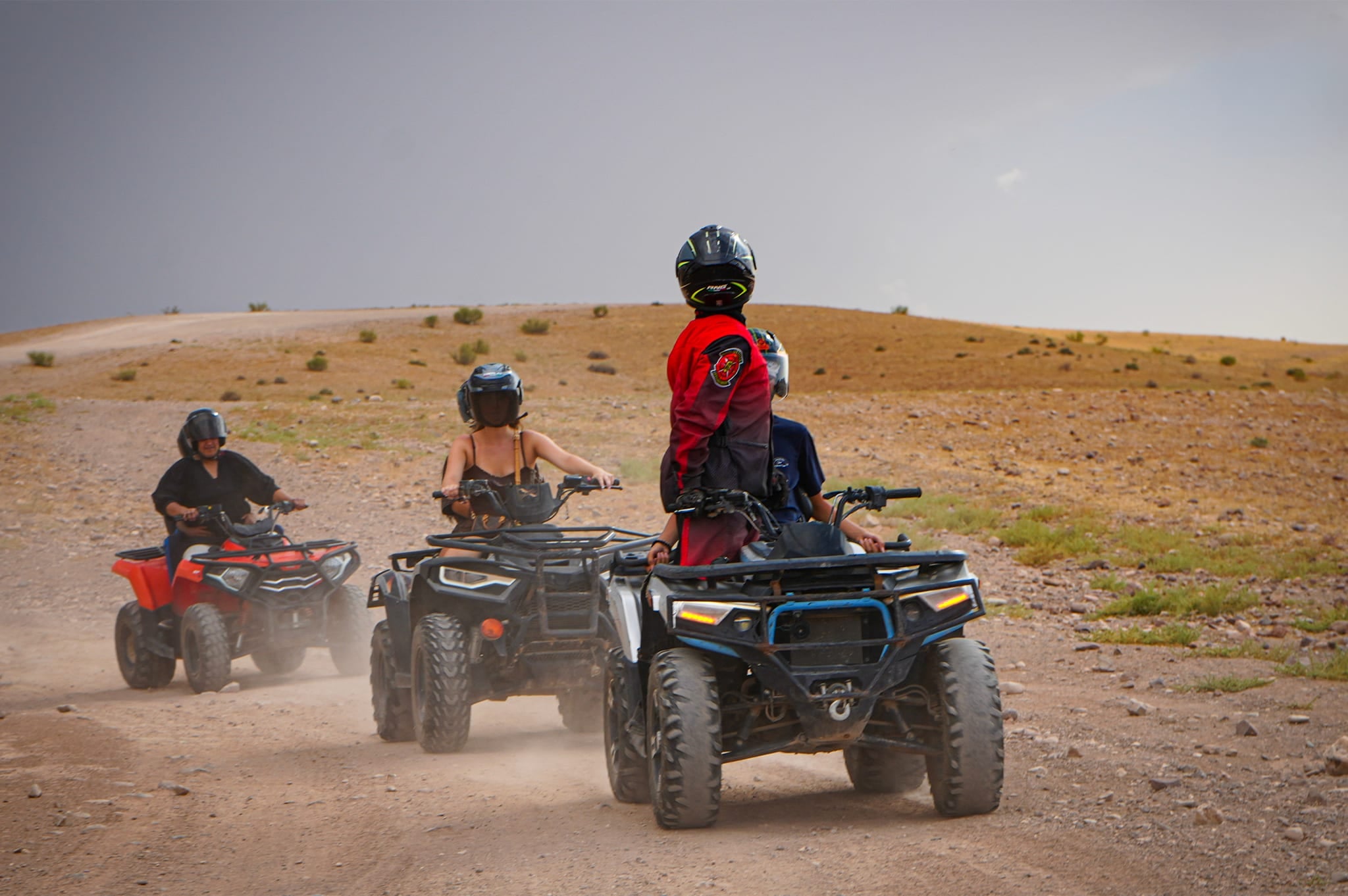 Pilota con la giacca rossa in piedi su un quad, girato verso i quad che lo seguono su una pista polverosa